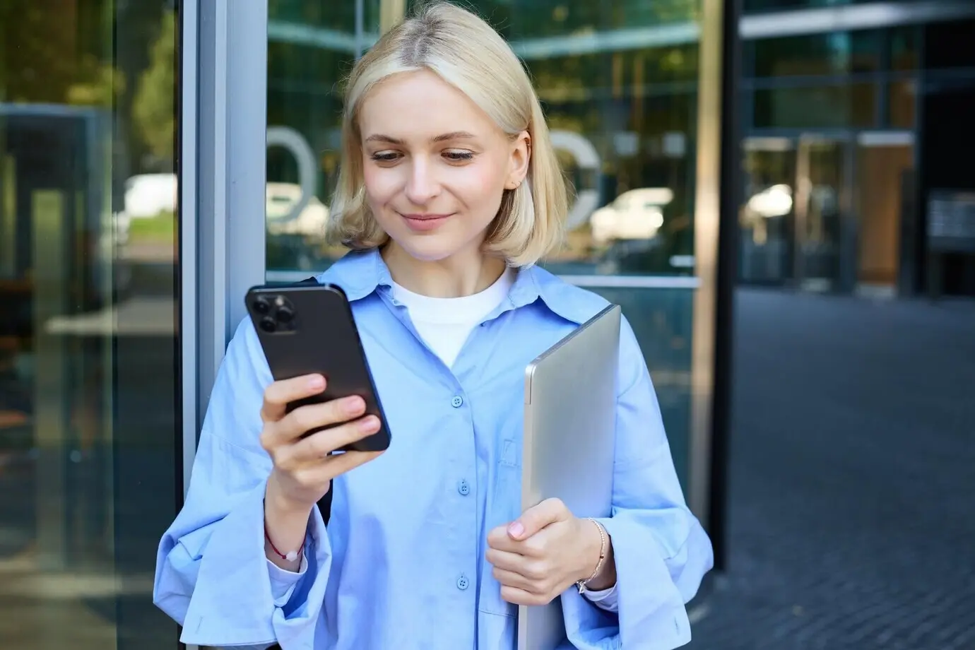 Fotografía de estilo de vida de una mujer joven sonriente, estudiante o empleada, cerca de un edificio de oficinas, sosteniendo un teléfono móvil.