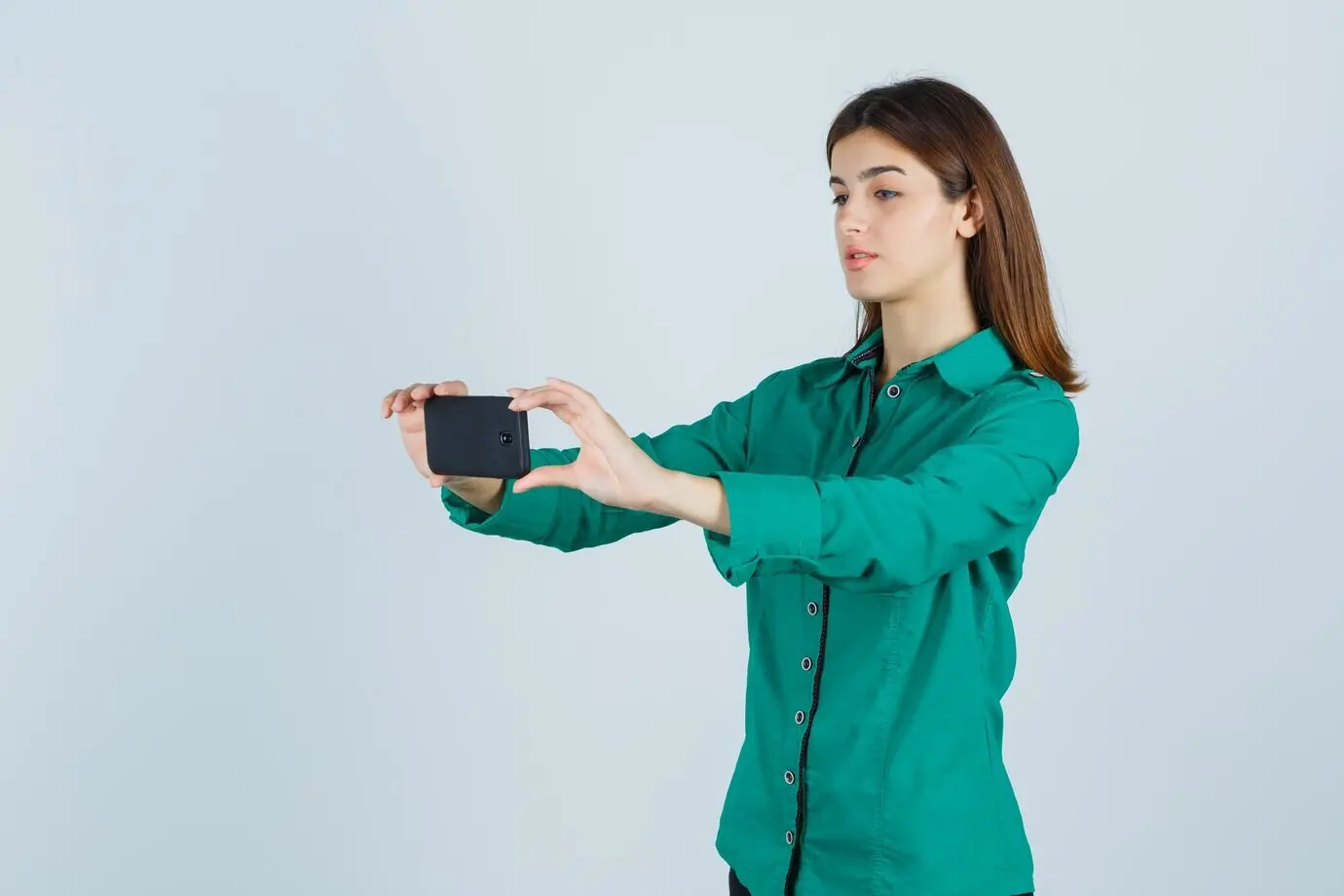 Chica joven tomándose una selfie con blusa verde, pantalones negros y expresión concentrada. Vista frontal.